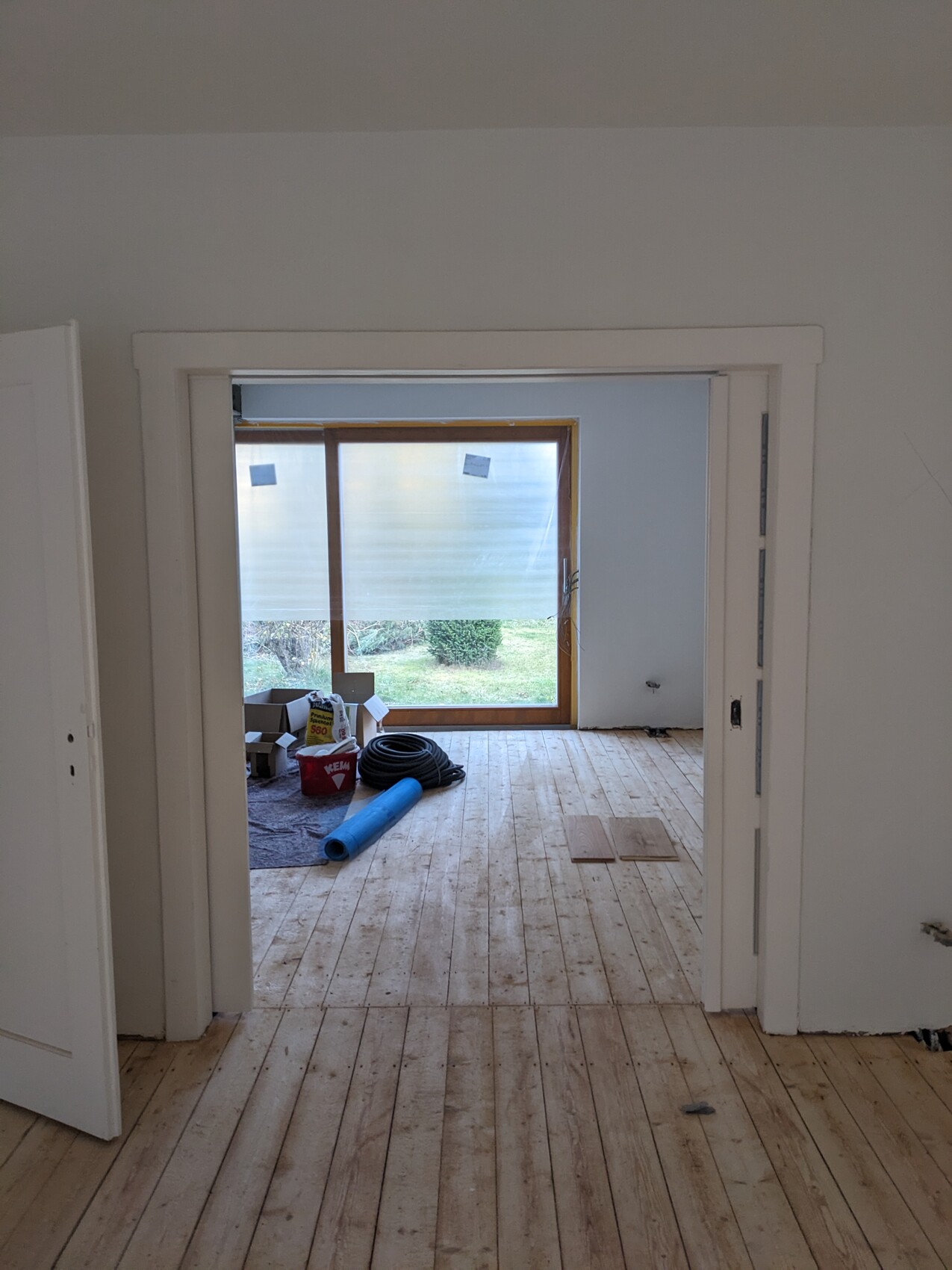 Rohbaustelle eines Zimmers mit Holzboden, Glasschiebetür zum Garten, Baumaterialien im Raum.