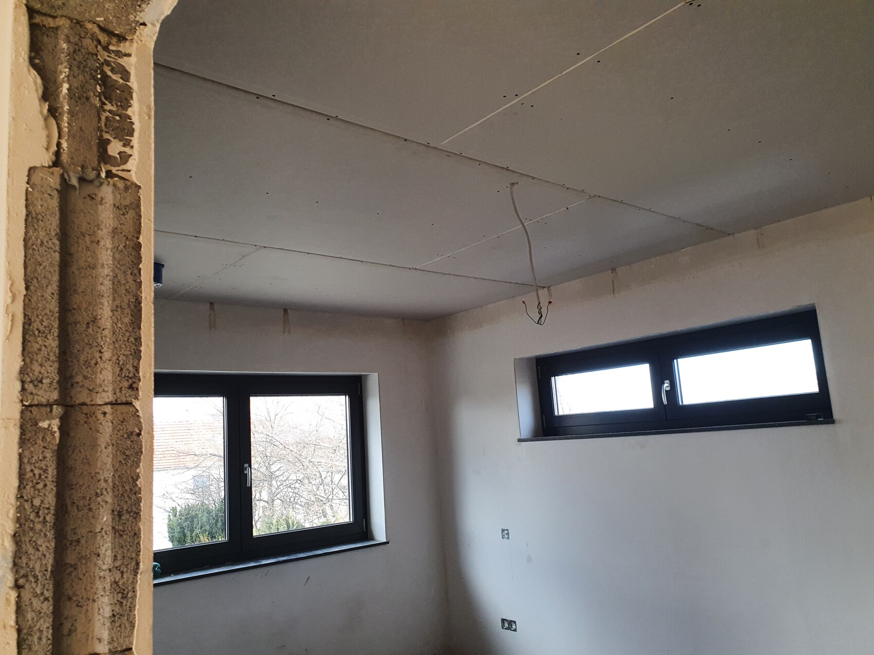 Rohbauzimmer mit zwei Fenstern, freiliegende Kabel, unfertige Wände.