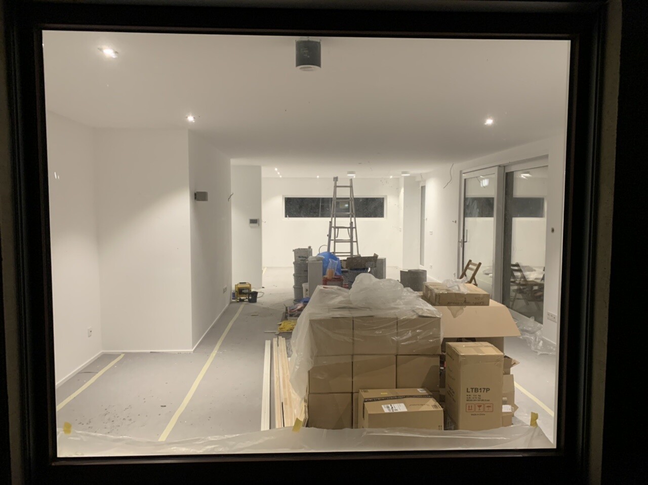 Innenraum einer Baustelle mit Leiter, Kartons, Plastikfolie und Glas-Schiebetüren rechts.