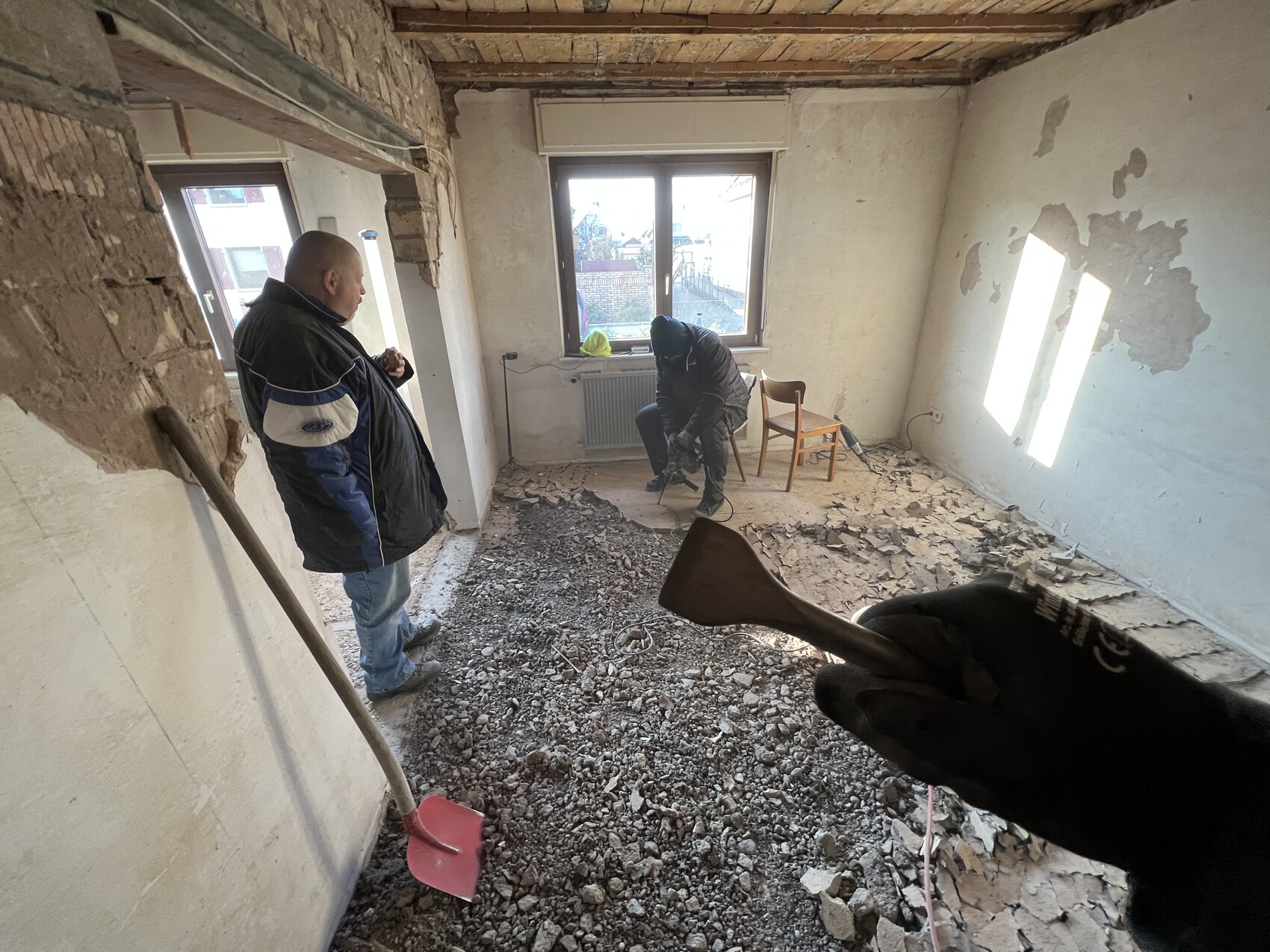 Zwei Bauarbeiter renovieren eine ruinöse Wohnung; Boden voller Schutt, einer arbeitet mit Werkzeug.