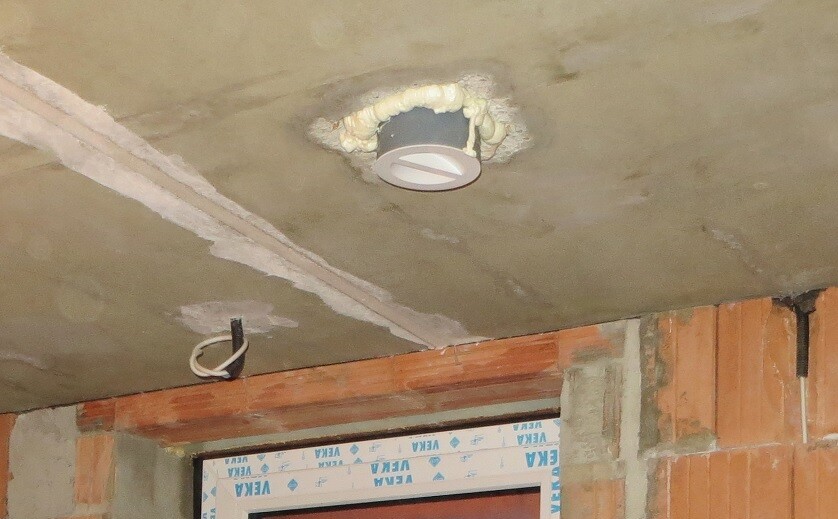 Runder Deckenspots in unfertiger Decke; gelber Bau-Schaum ringsum, lose Kabel, Ziegelwand sichtbar.
