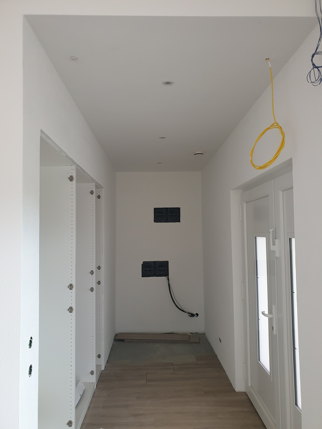 Heller Bau-Flur: weiße Wände, offene Schränke links, gelbes Kabel von der Decke, Tür mit Glas rechts