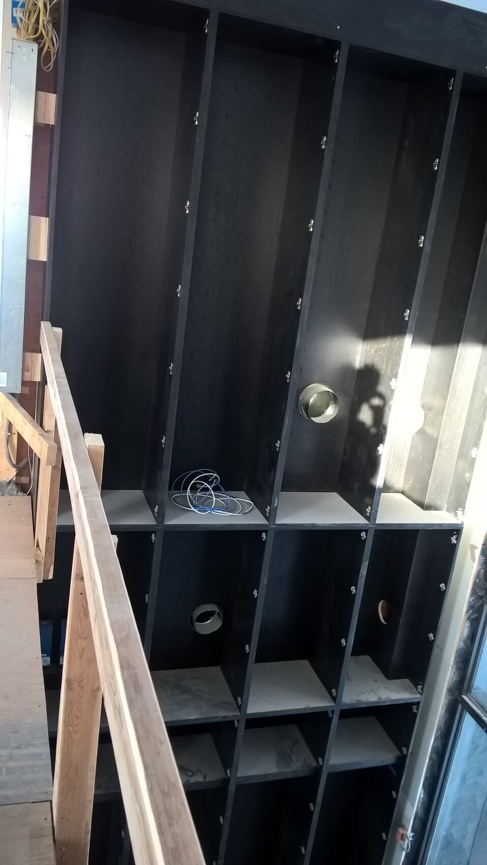 Schwarze Regale mit Fächern, runde Rohröffnungen und lose Kabel; Holztreppe links.