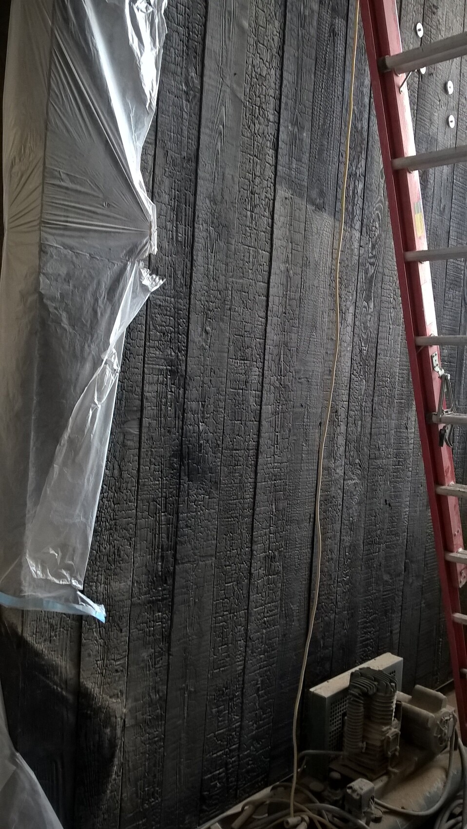 Verbrannte schwarze Holzwand mit roter Leiter davor; Plastikfolie links, Maschine unten rechts