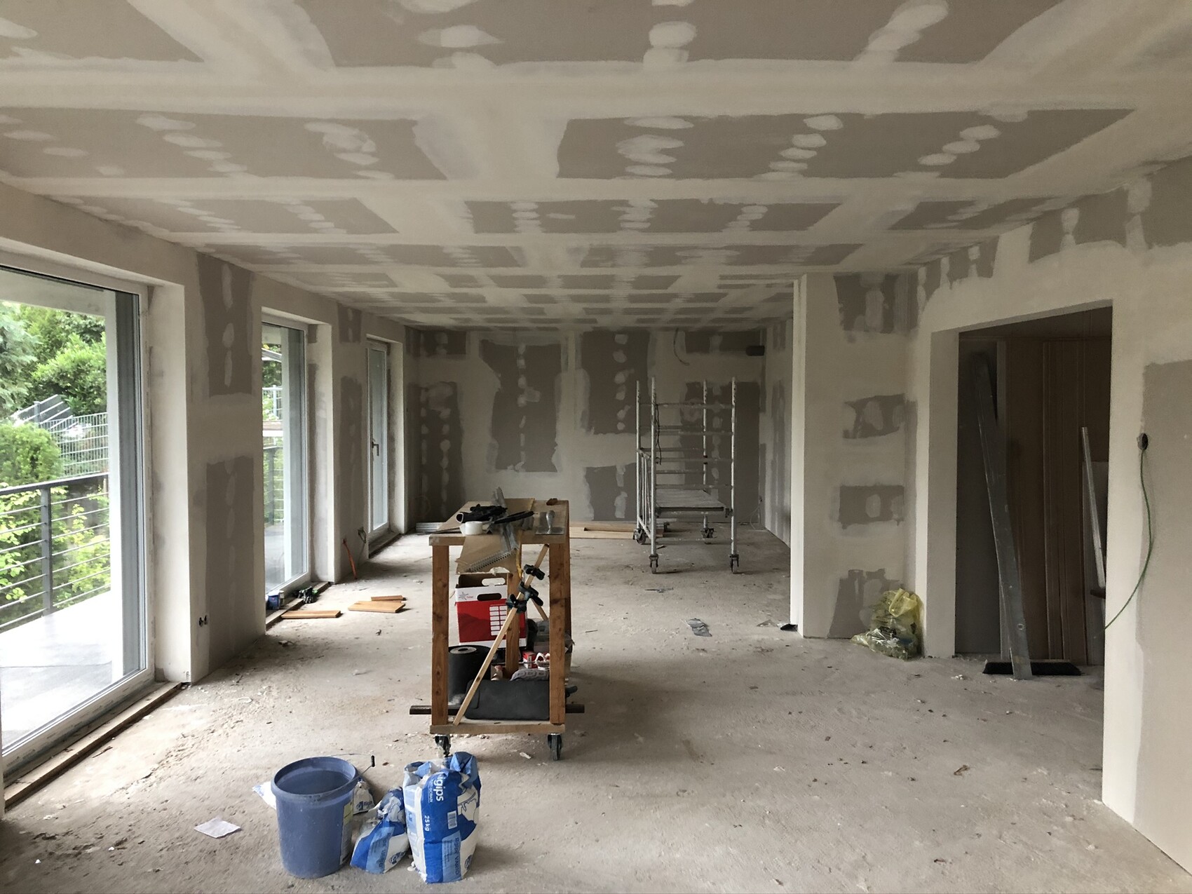 Rohbau eines Wohnzimmers mit verputzten Wänden, Werkzeugen, Gerüst und großen Fenstern.