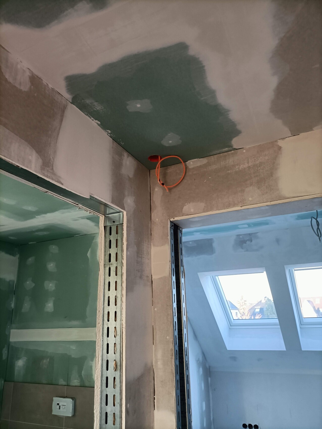 Baustelle: unfertige Wände, Metallständer, rotes Kabel von der Decke, Dachfenster im Hintergrund.