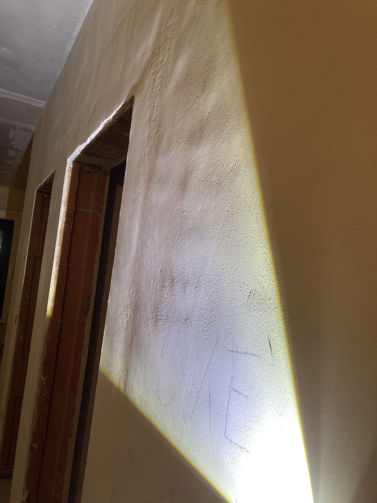 Innenraum: rauhe Wand mit offenen Türrahmen aus Backstein; gelber Lichtstreifen trifft die Wand.
