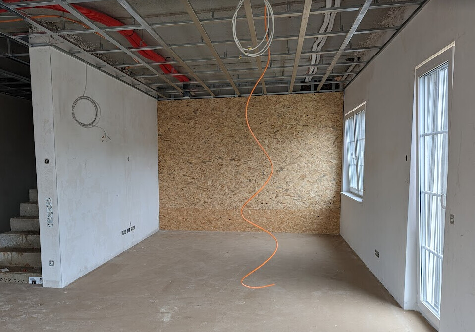 Innenraum im Bau: OSB-Wand, freigelegter Deckenbereich, orange Kabel hängt herab, Fenster rechts.