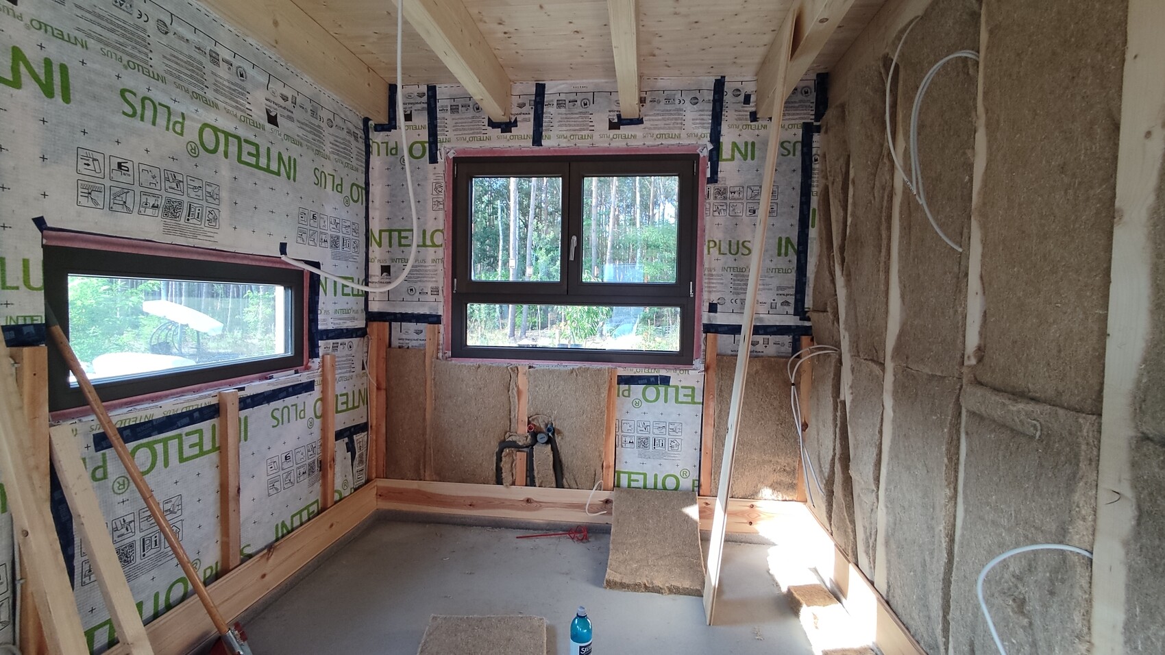 Unfertiger Innenraum eines Hauses mit Holzständern, Dämmung, zwei Fenster, Kabeln und Baumaterial.