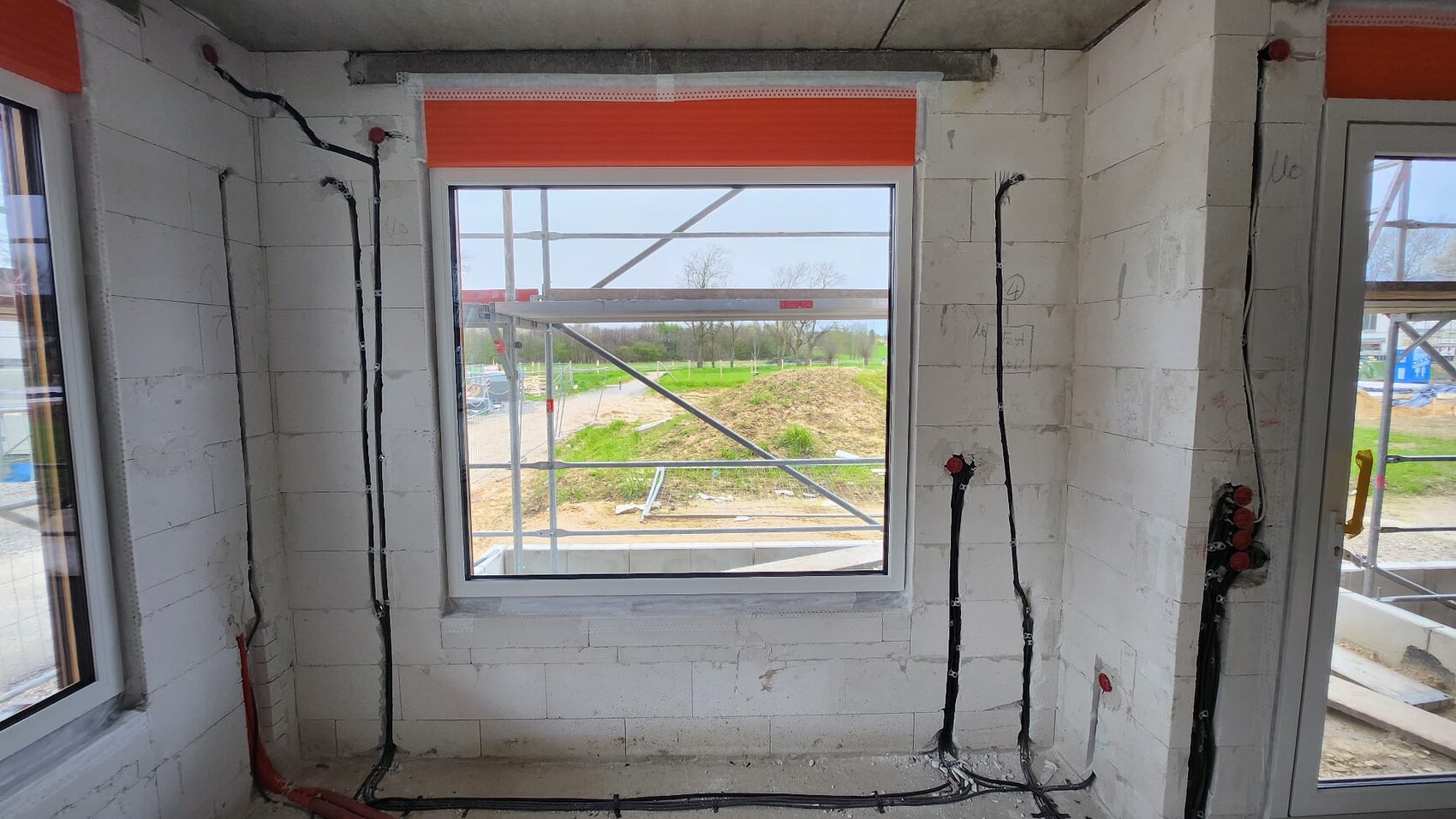 Innenraum eines Bauprojekts mit freiliegenden Kabeln, roten Abdeckkappen und Fensterblick auf Gerüst.