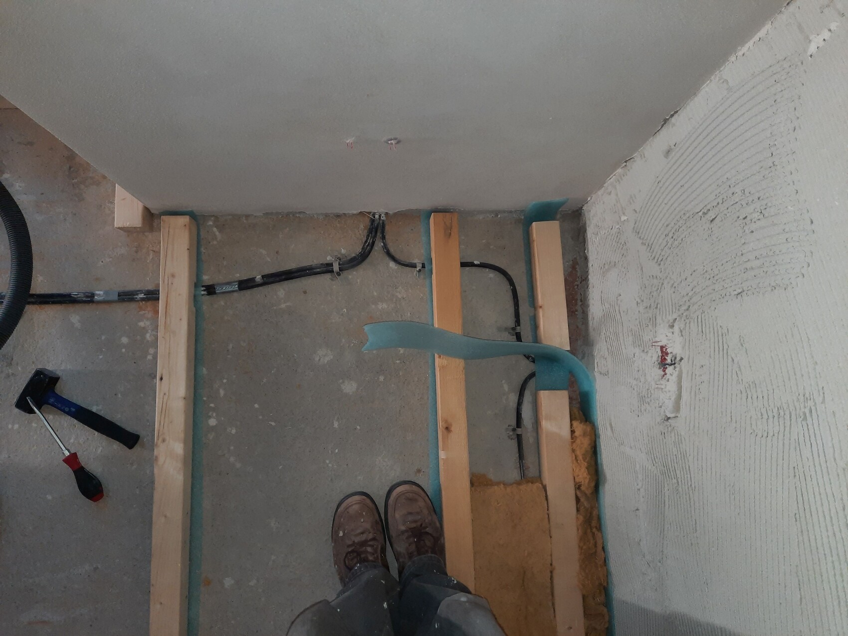 Rohbau: Holzstaender-Wand, blaue Daemmplatten, schwarze Kabel, Hammer links, Schuhe unten.