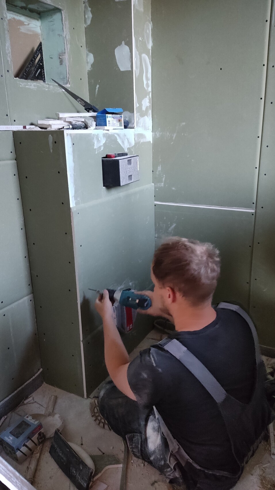 Bauarbeiter kniet vor grünem Trockenbau und bohrt mit Akku-Bohrer in der Wand.