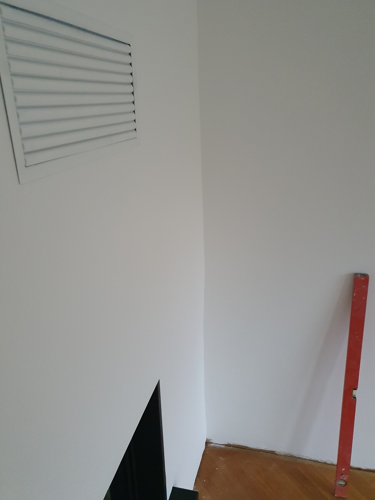 Weiße Innenwand mit Lüftungsgitter, rote Wasserwaage an der Wand, Holzboden sichtbar.