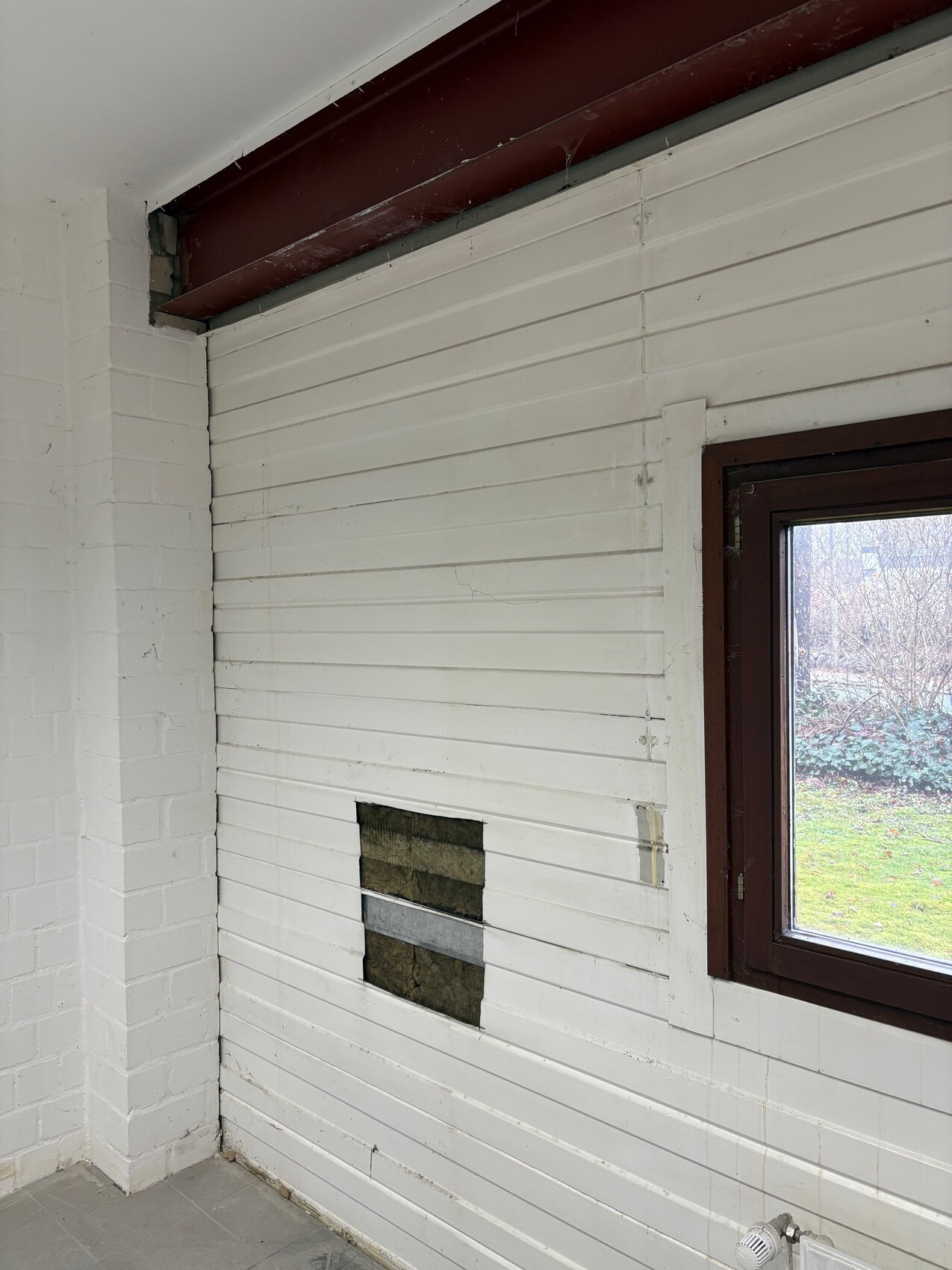 Weiße Wand mit horizontalen Paneelen, brauner Fensterrahmen und einer rechteckigen Öffnung mit Dämmmaterial.