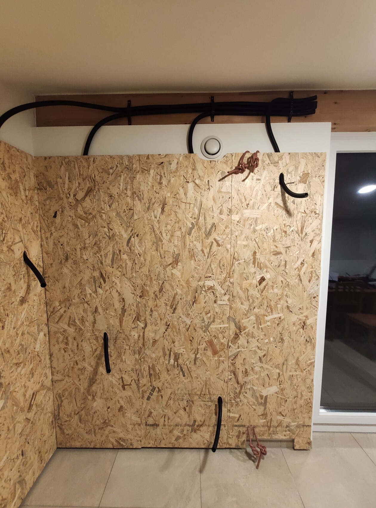 OSB-Plattenwand in einem Innenraum, lose schwarze Kabel und Schläuche hängen an Decke und Wand.