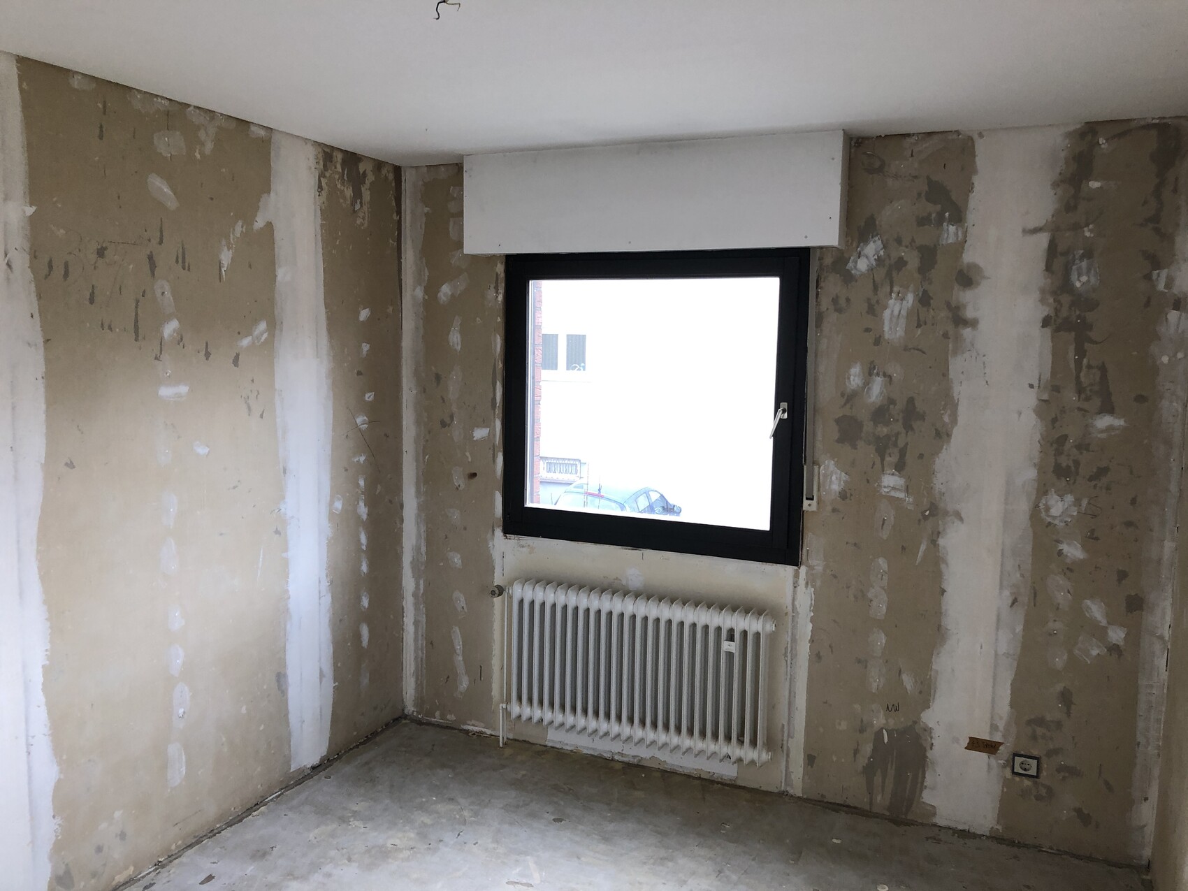 Unfertiges Innenraumzimmer mit rohen Wänden, Fenster und Heizkörper darunter.