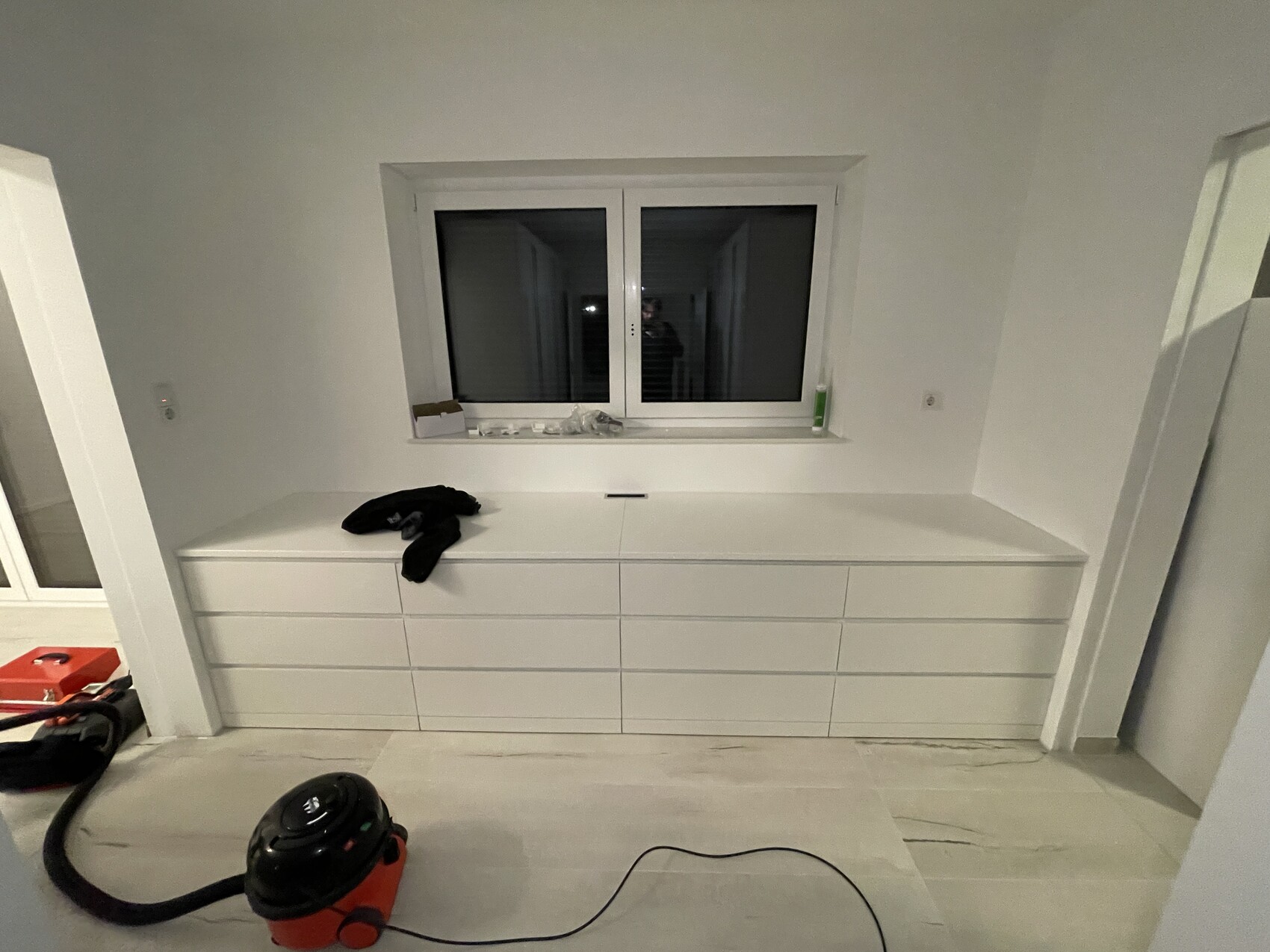 Weiße, moderne Kommode unter Fenster; schwarzes Kleidungsstück liegt darauf, Staubsauger am Boden.