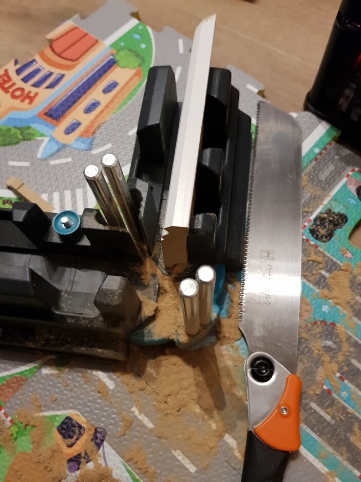 Arbeitsplatte mit Sägeführung: Handsäge mit orangefarbenem Griff, Metallstifte in Halterung und Holzblock, Sägespäne.