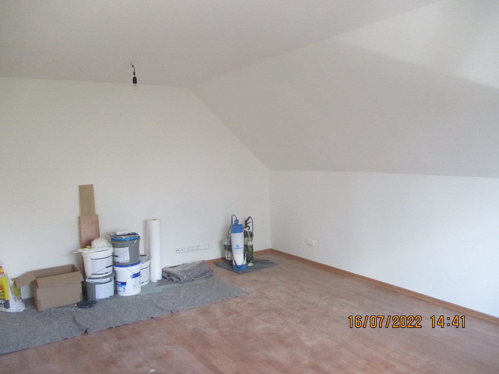 Renovierungsraum mit schräger Decke; links Farbdosen, Kartons und Abdeckmaterial.