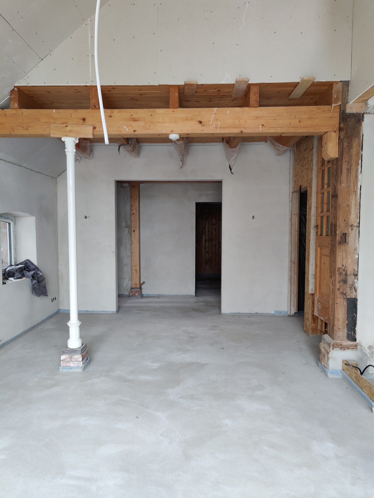 Offener Innenraum im Bau: Holzbalkendecke, weiße Stütze, rohe Wände, Türöffnungen.