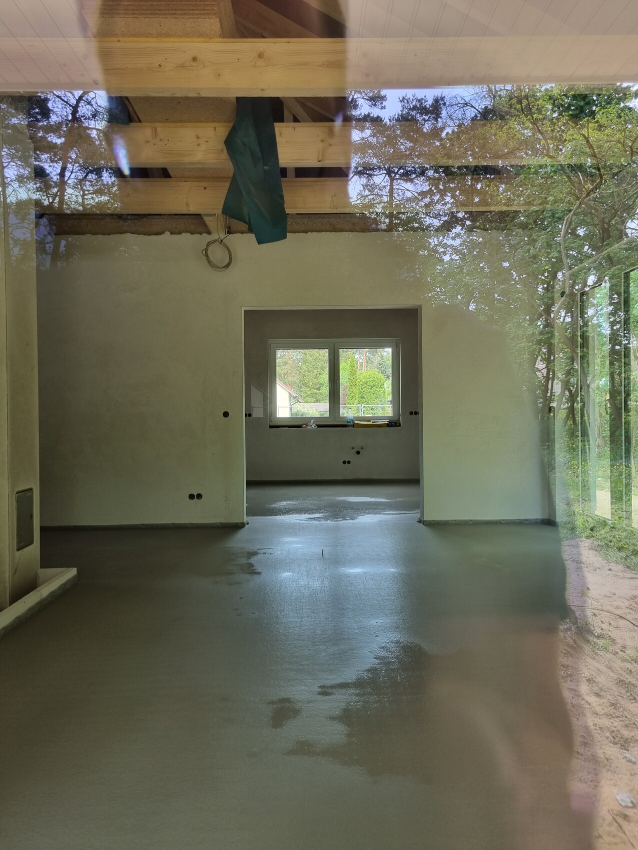 Renovierungsraum: offener Bereich mit Holzbalkendecke, feuchtem Boden und Fensterblick ins Freie.