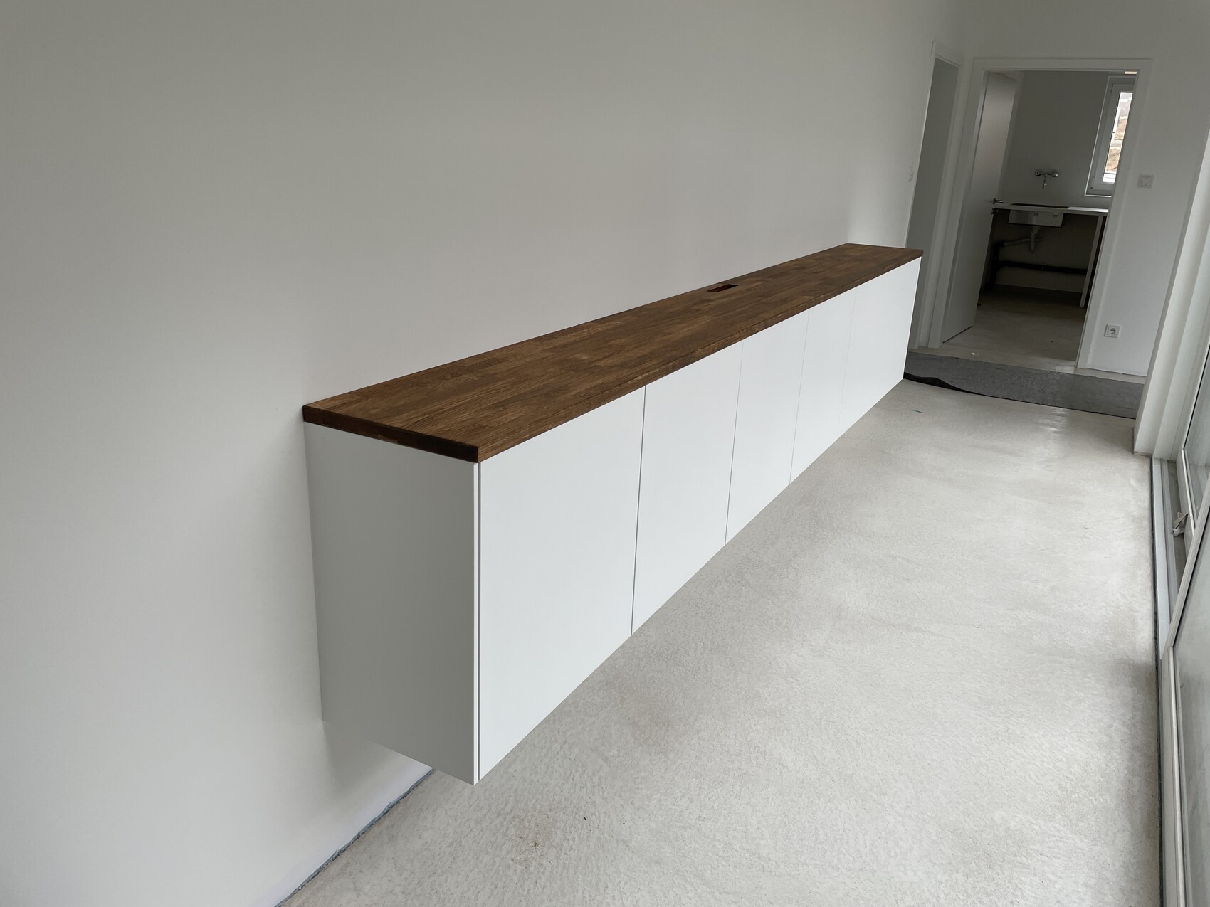 Langes weißes wandmontiertes Sideboard mit dunklem Holzoberteil vor heller Wand.