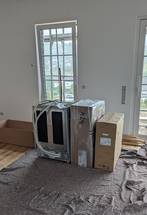 Unfertiger Innenraum mit verpackten Küchengeräten, Kartons und Holzlatten neben Fenstern.