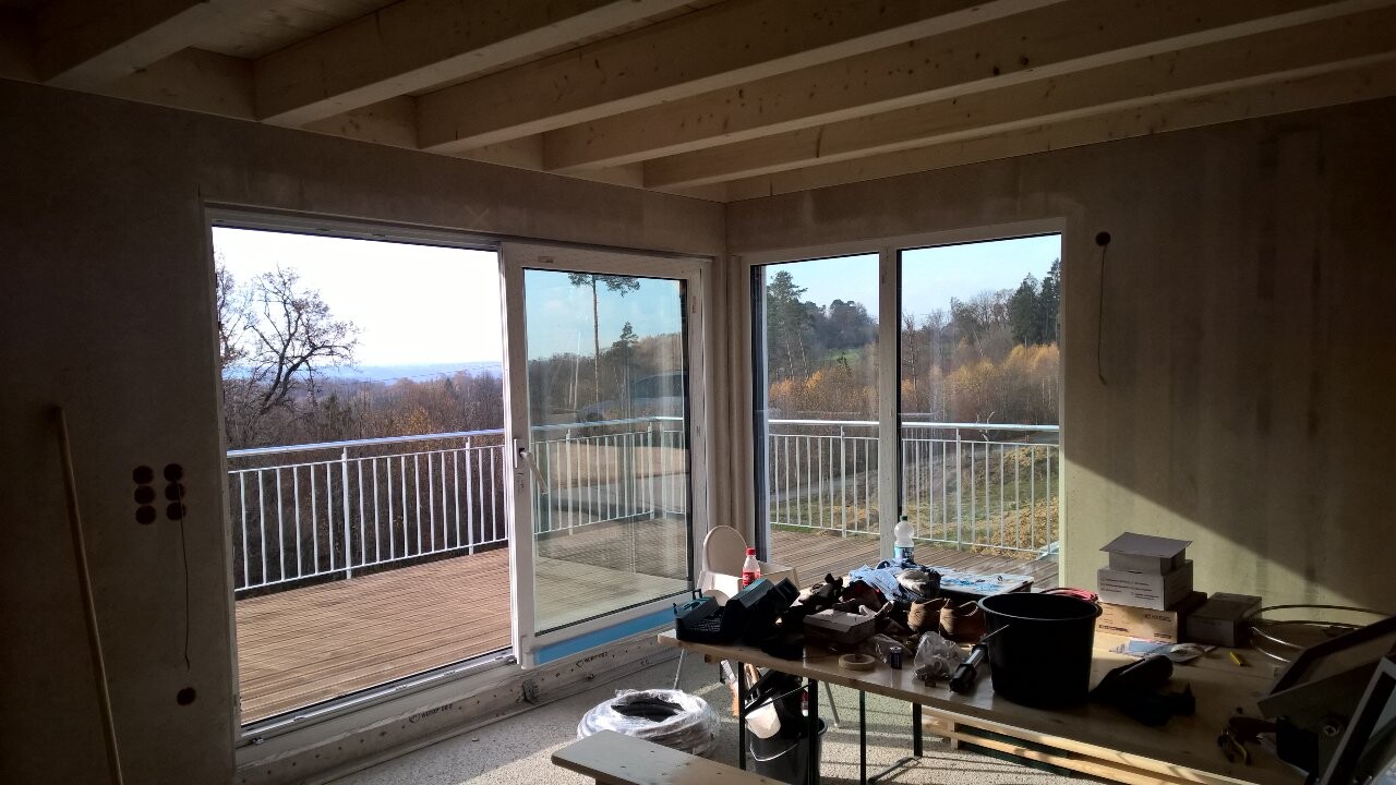 Innenraum im Rohbau mit großen Glas-Schiebetüren zur Terrasse; Bauwerkzeuge auf Tisch.