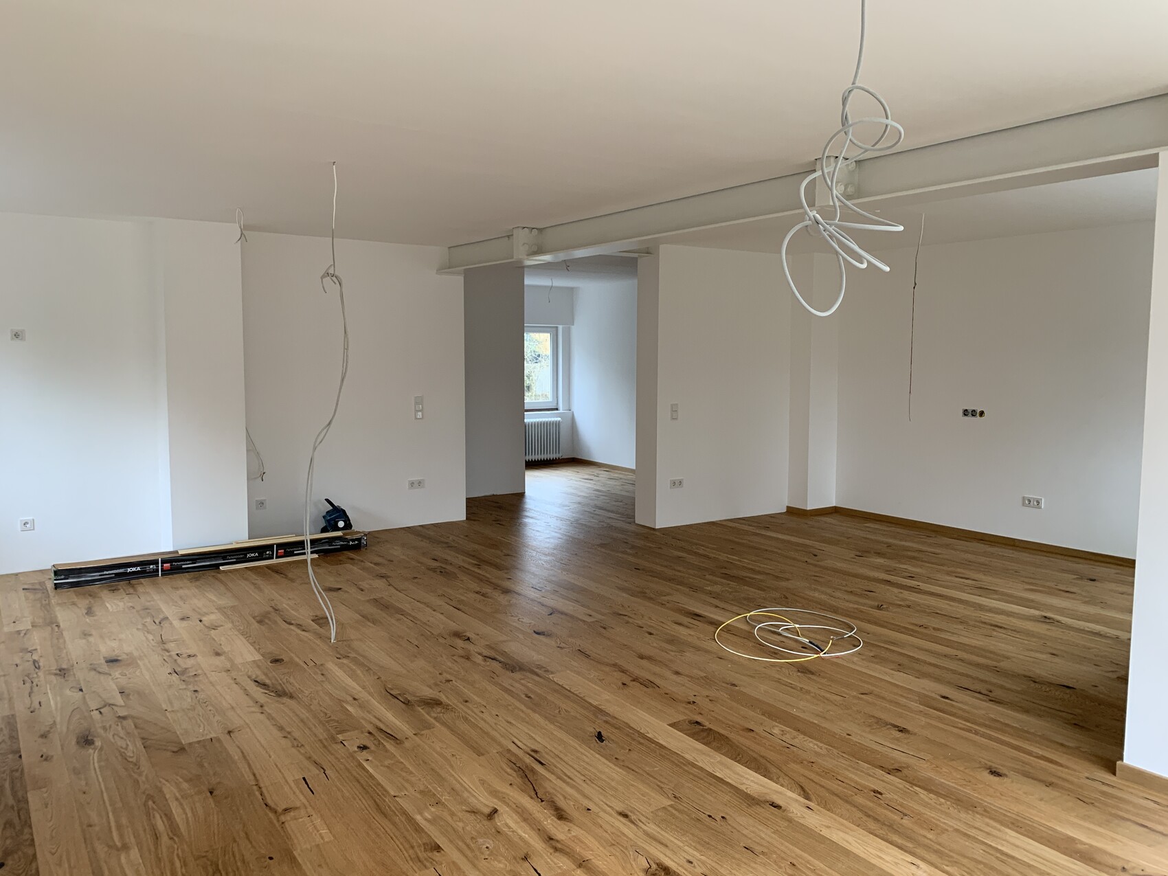 Leeres, renoviertes Wohnzimmer mit Holzboden; hängende Kabel von der Decke, Werkzeug auf dem Boden.