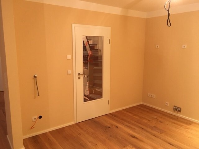 Leerer Raum mit Holzboden, beige Wände, weiße Tür mit Glaseinsatz, lose Kabel.