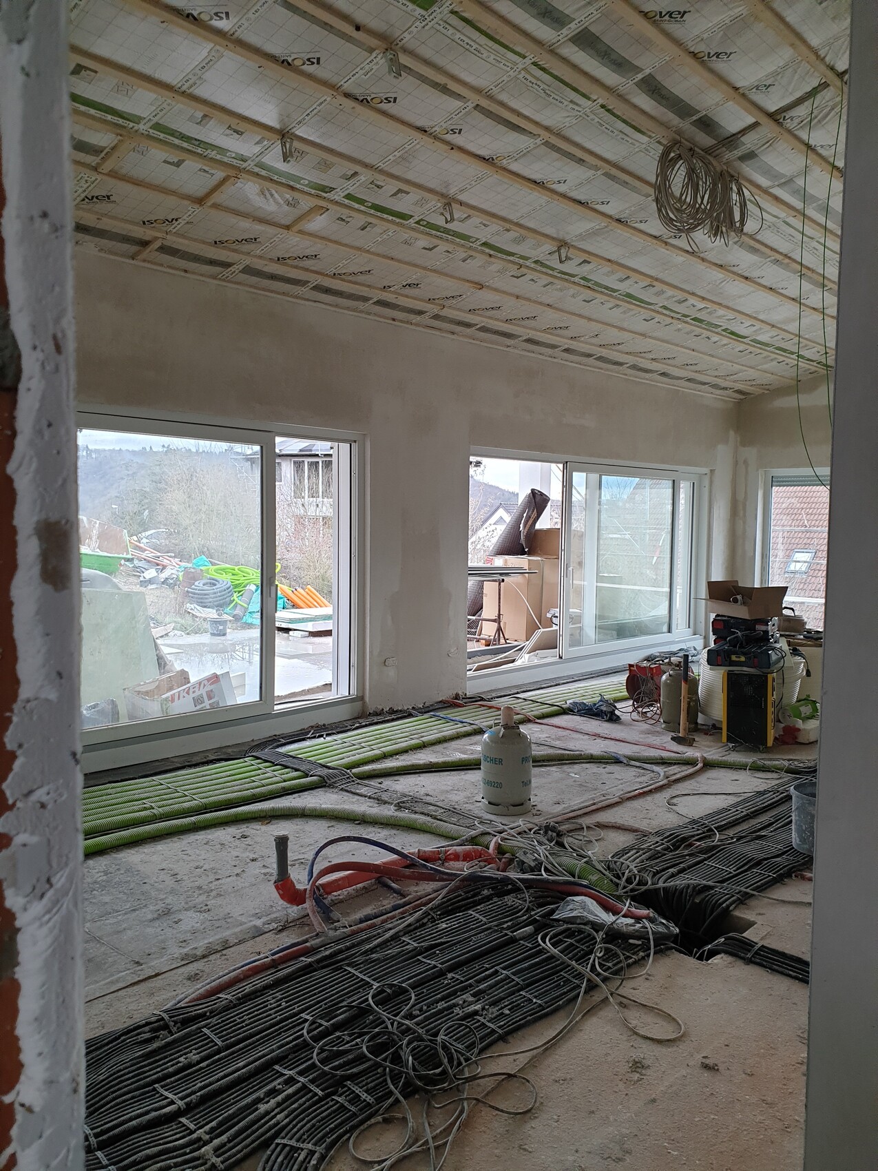 Innenraum einer Baustelle: Deckenisolierung, Kabel, Rohre, Fensterfront, Bauwerkzeuge.