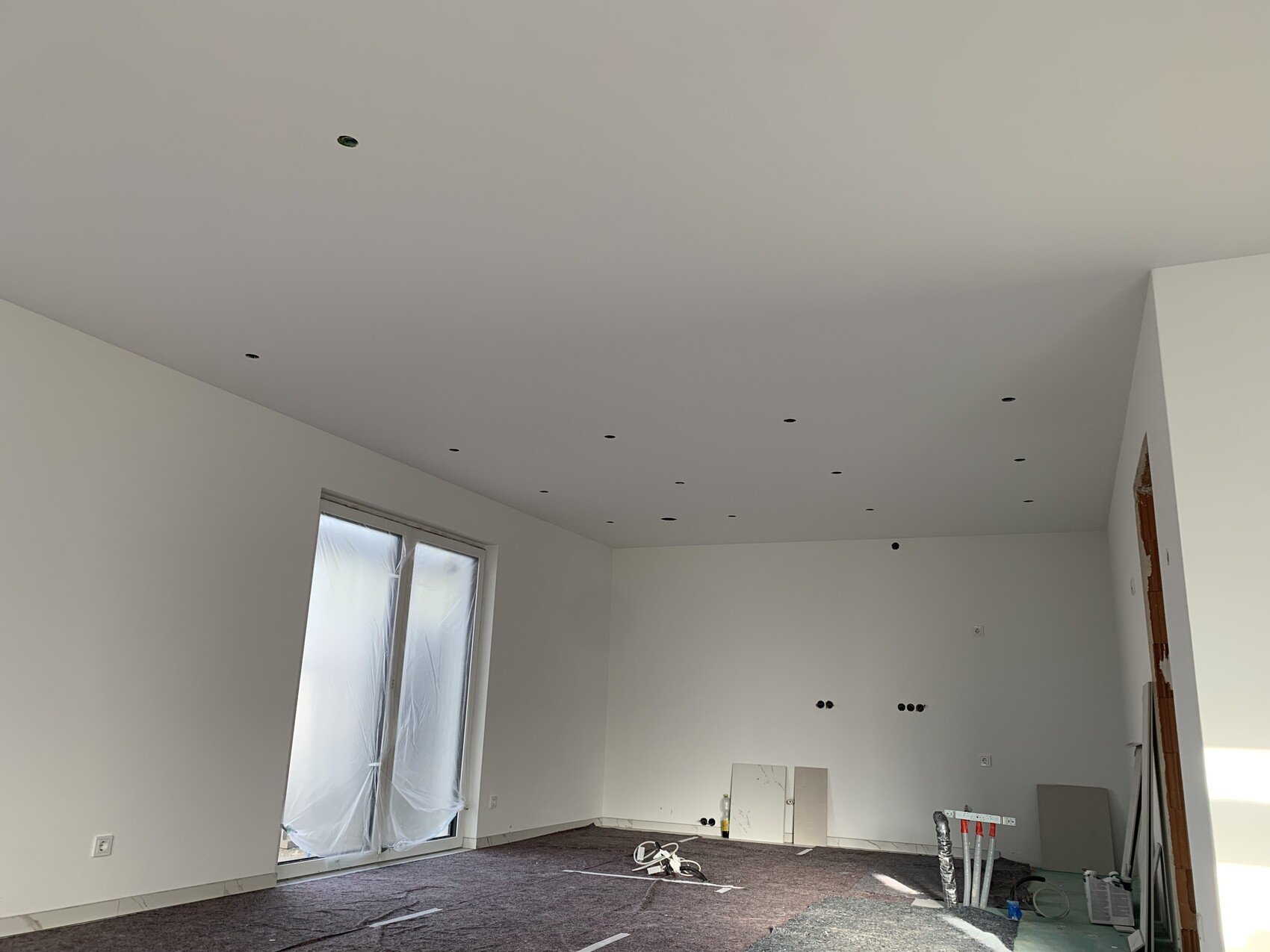 Leerer Bau-Wohnraum mit weißen Wänden, Deckenlöcher, verpackte Glasfront und Baustoffe