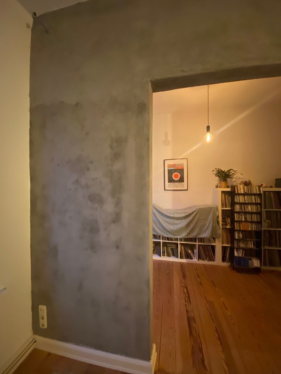 Wohnzimmer mit rohem Betonwandabschnitt, Holzfussboden, Buecherregale, Haengelampe.