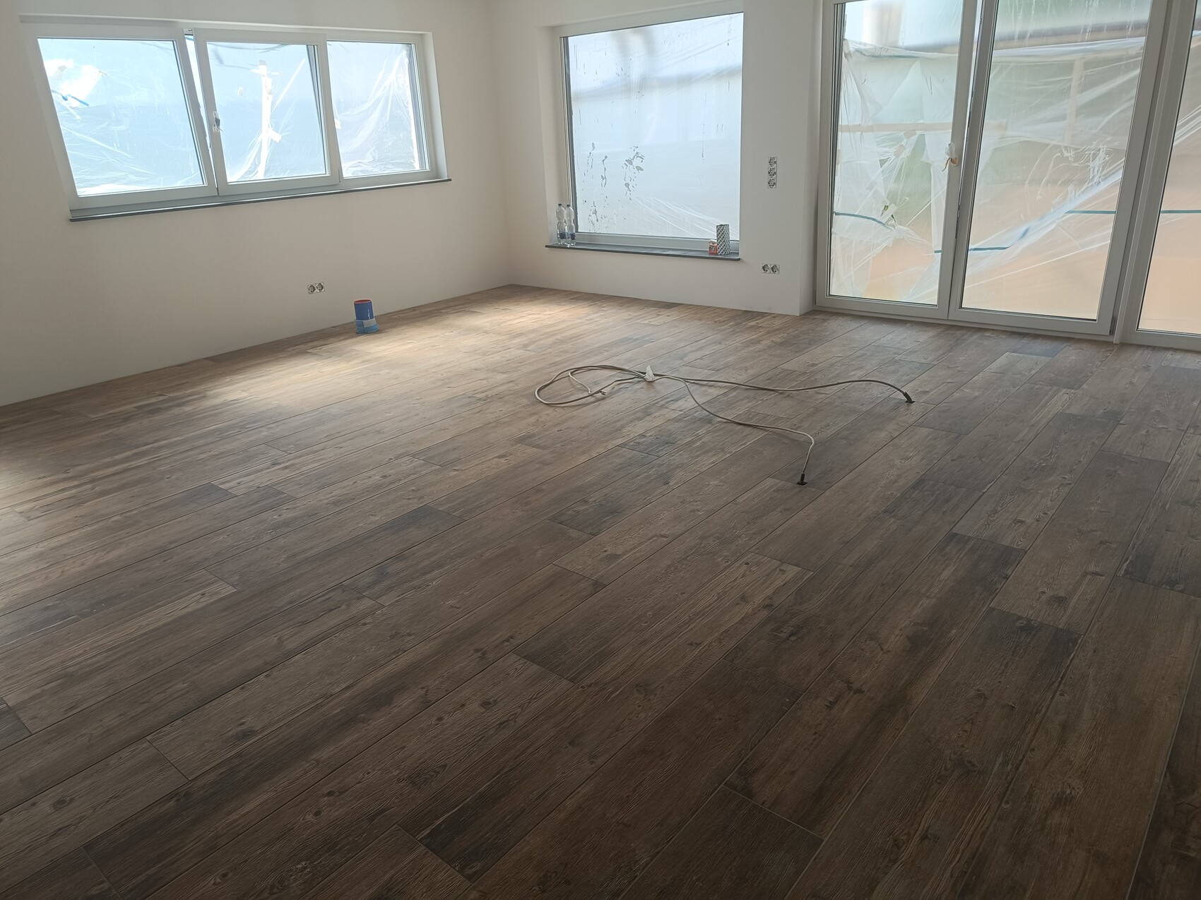 Heller leerer Raum mit Holzlaminatboden; Fensterfront hinter Plastikfolie; Kabel auf dem Boden