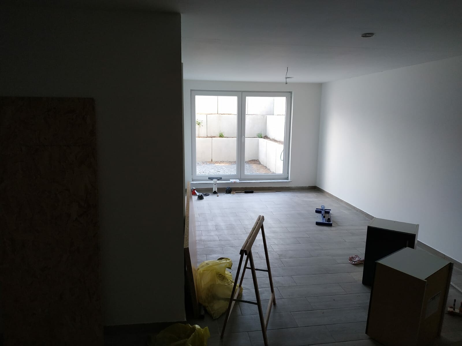 Renovierungsarbeiten in einem leeren Raum: große Fenstertür zur Terrasse, Werkzeug, Leiter und Kartons auf dem Boden.