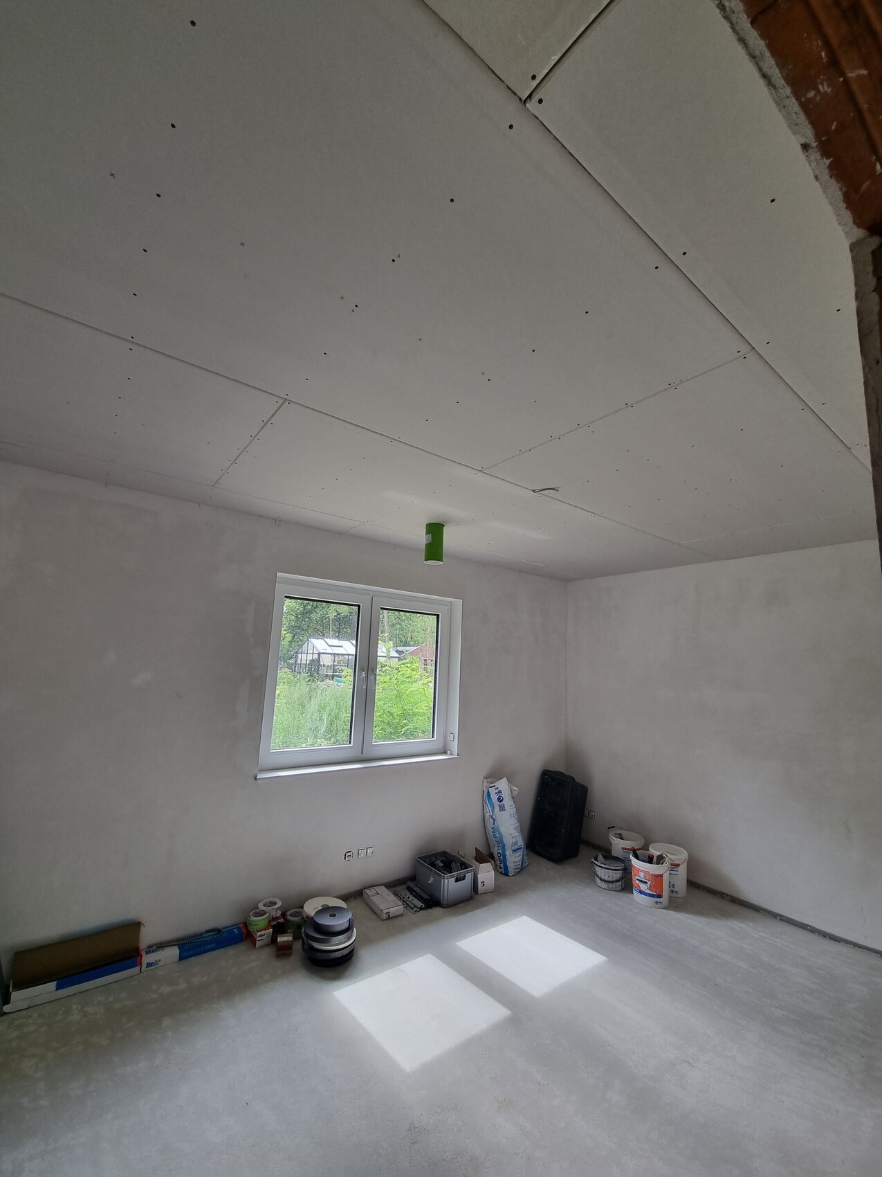 Unfertiger Raum mit Trockenbau-Decke, Fenster und Baumaterialien am Boden.