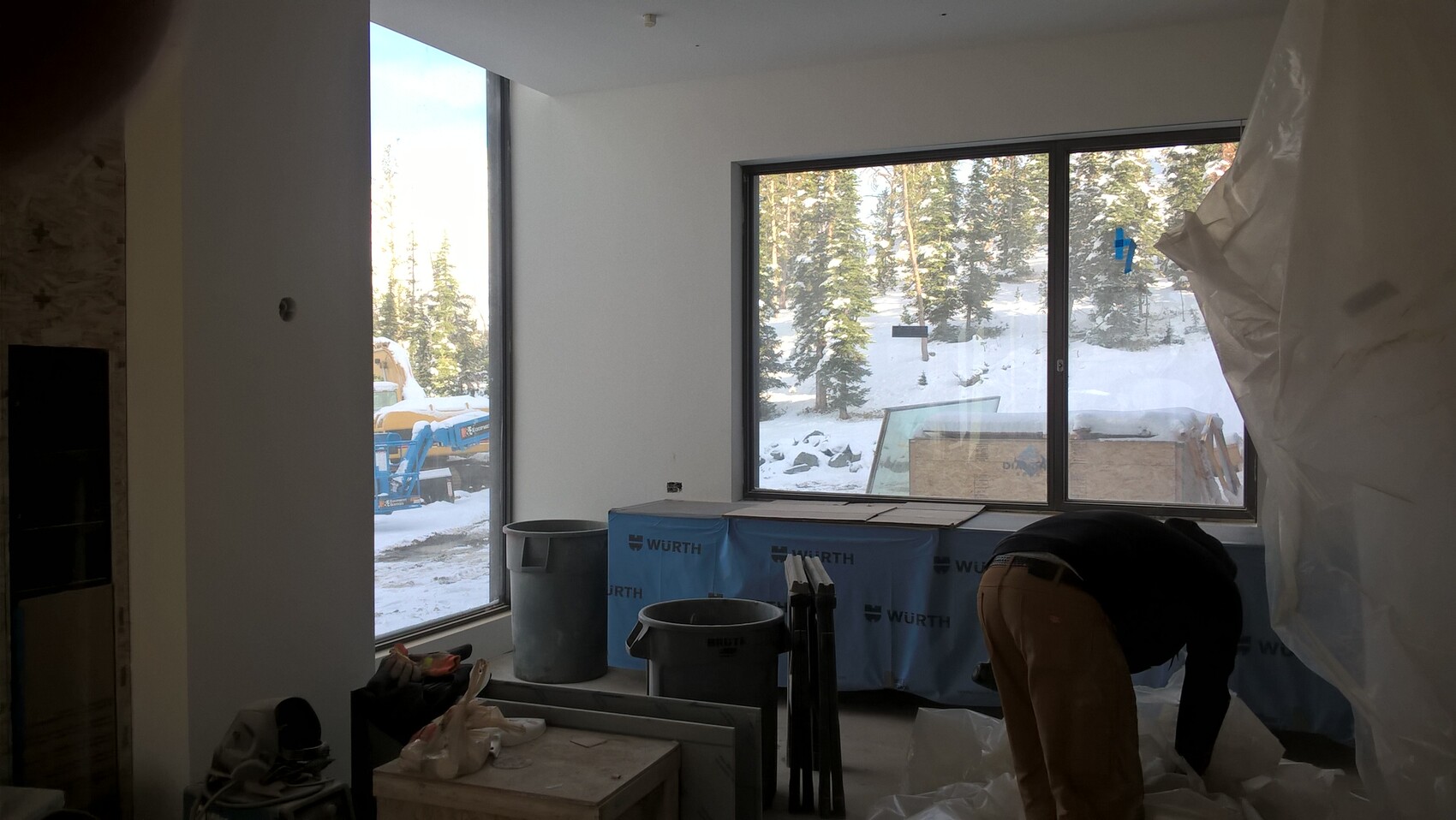 Baustelle im Innenraum: große Fenster, Schnee draußen, Werkzeuge und eine Person beim Arbeiten.