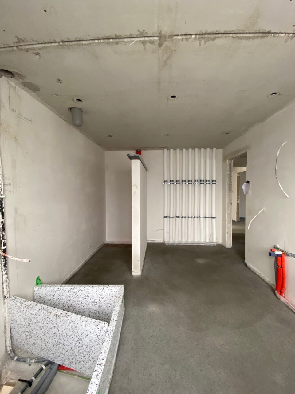 Baustellenraum: Rohbau mit weißen Sanitärrohren an der Wand, Stütze in Mitte, Treppenstufen links.