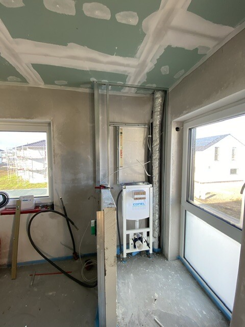 Innenraum einer Baustelle mit Heizgerät, Rohren, Elektroinstallationen und unfertigem Trockenbau.