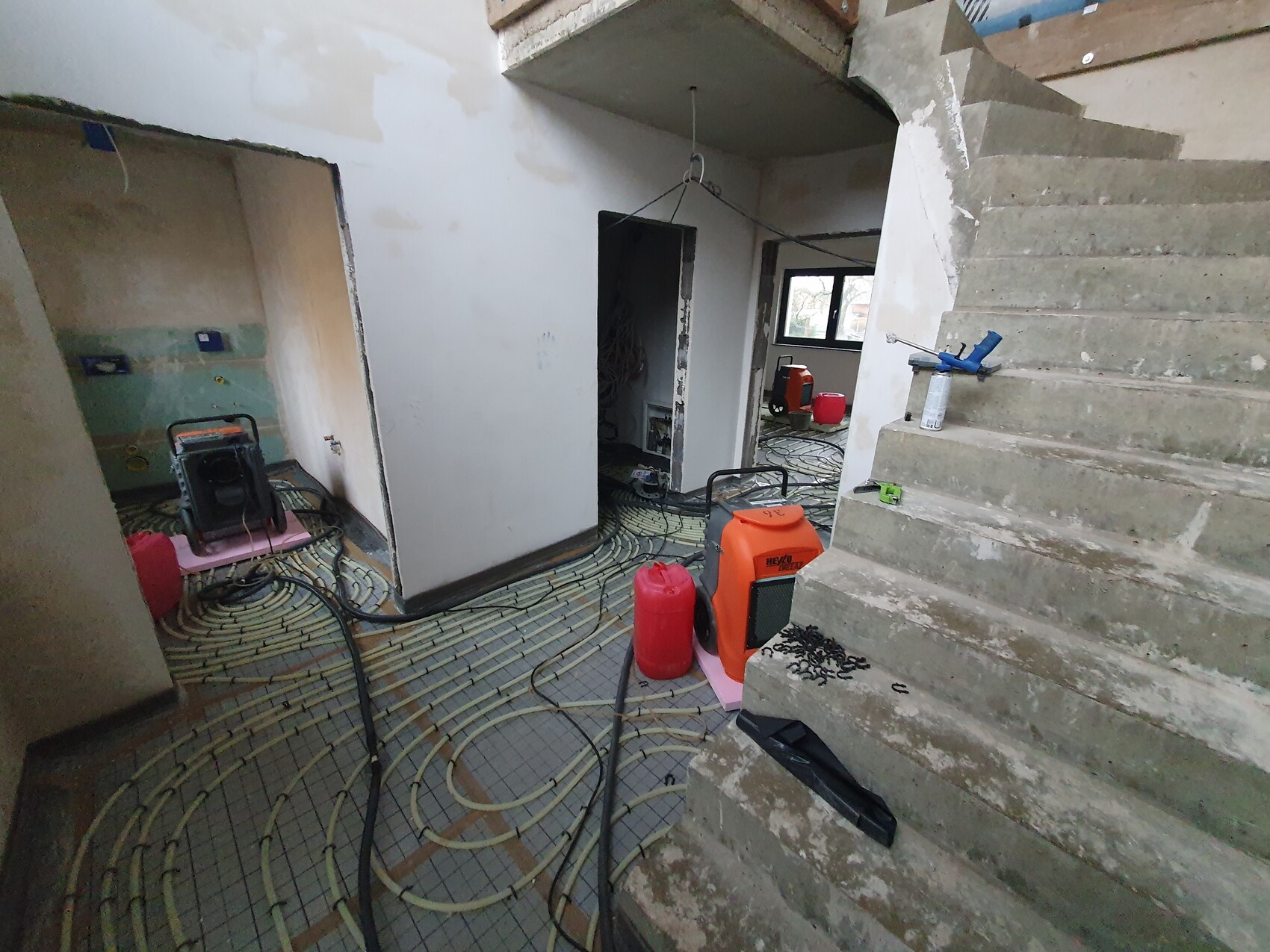 Innenraum-Baustelle: verlegte Heizungsrohre auf dem Boden, Betonstufen rechts, Werkzeuge.