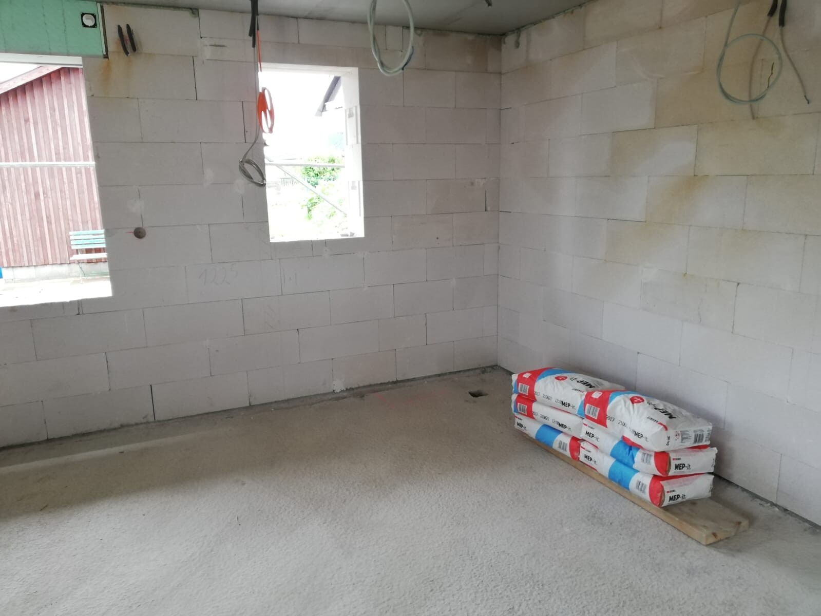 Unfertiger Innenraum mit weißen Blöcken, Hängekabeln, Fensteröffnung und Baustoffpaketen am Boden.