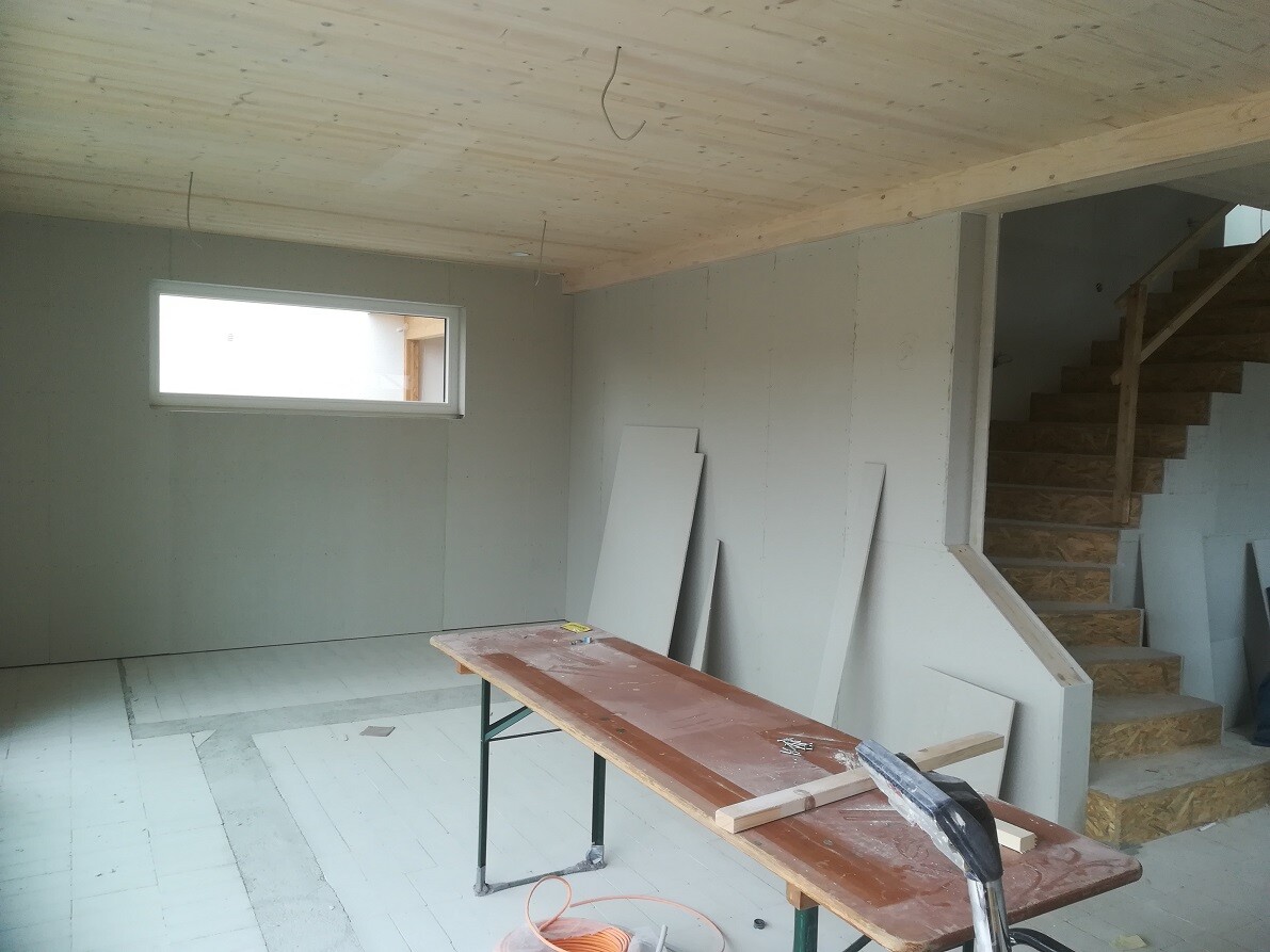 Raum im Rohbau mit Holzdecke, kleinem Fenster, Treppe und Arbeitsbank mit Baumaterial.