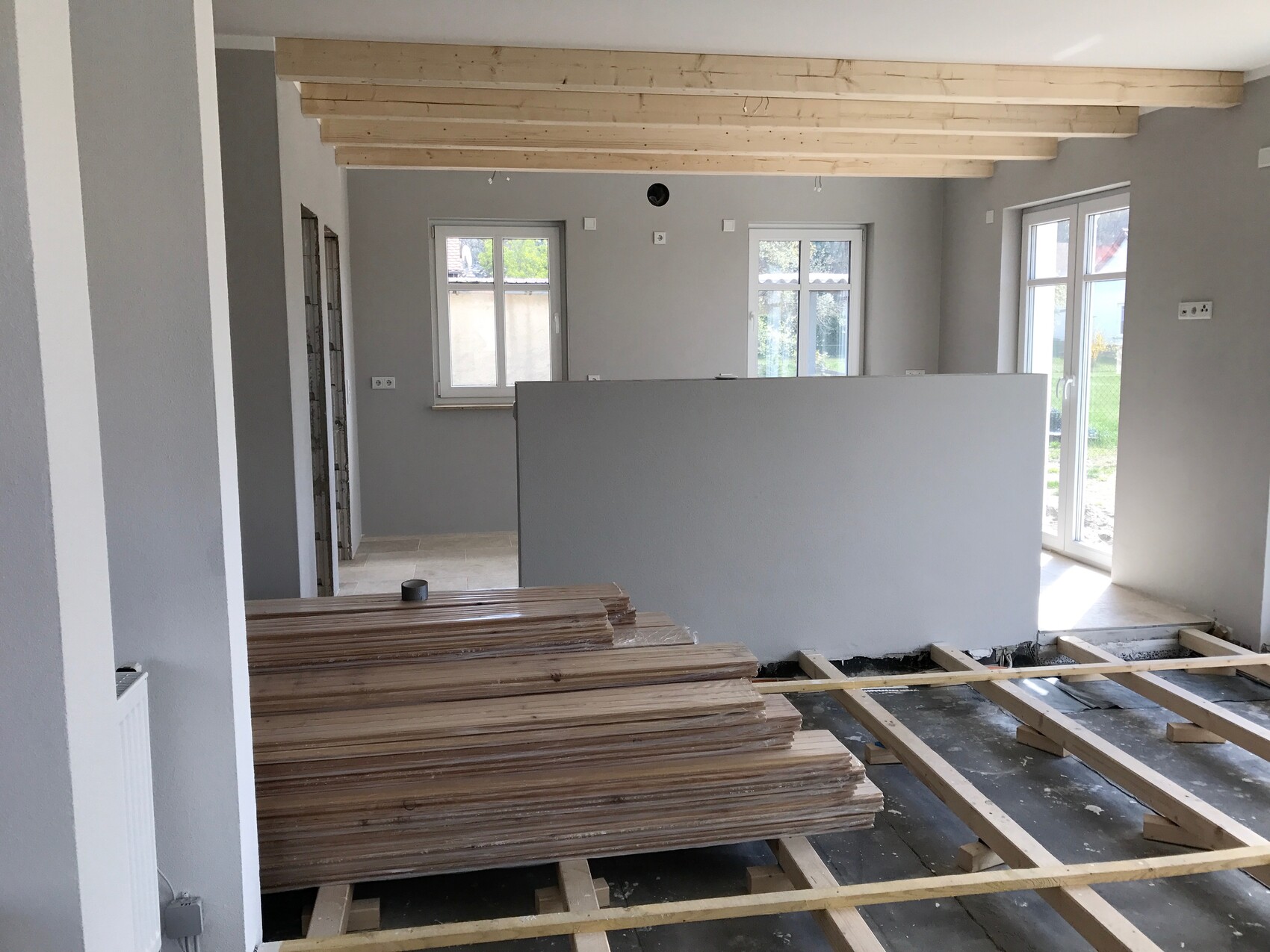 Rohbau eines Wohnraums: sichtbare Holzdeckenbalken, Trennwand, Fenster und gestapelte Holzbretter.