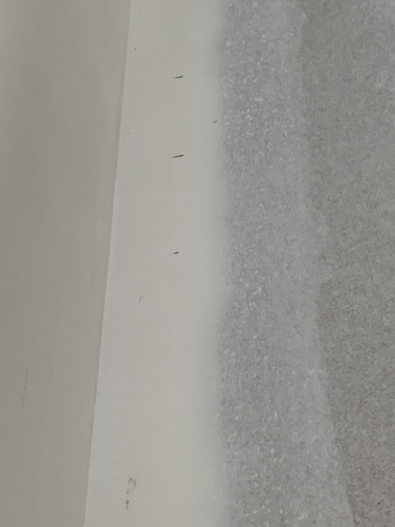 Weiße Wandkante mit kleinen dunklen Nägeln; rechts grauer, rauer Untergrund.