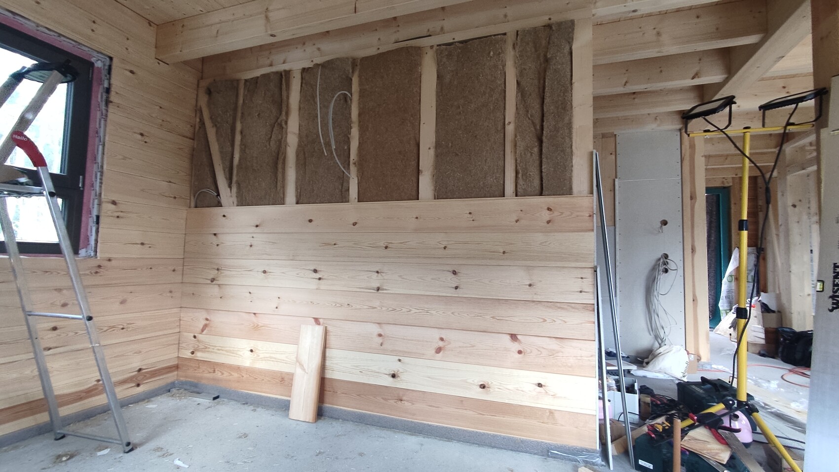 Innenraum eines Rohbaus: Holzverkleidung an Wänden, sichtbare Dämmung, Leiter und Bauwerkzeuge.