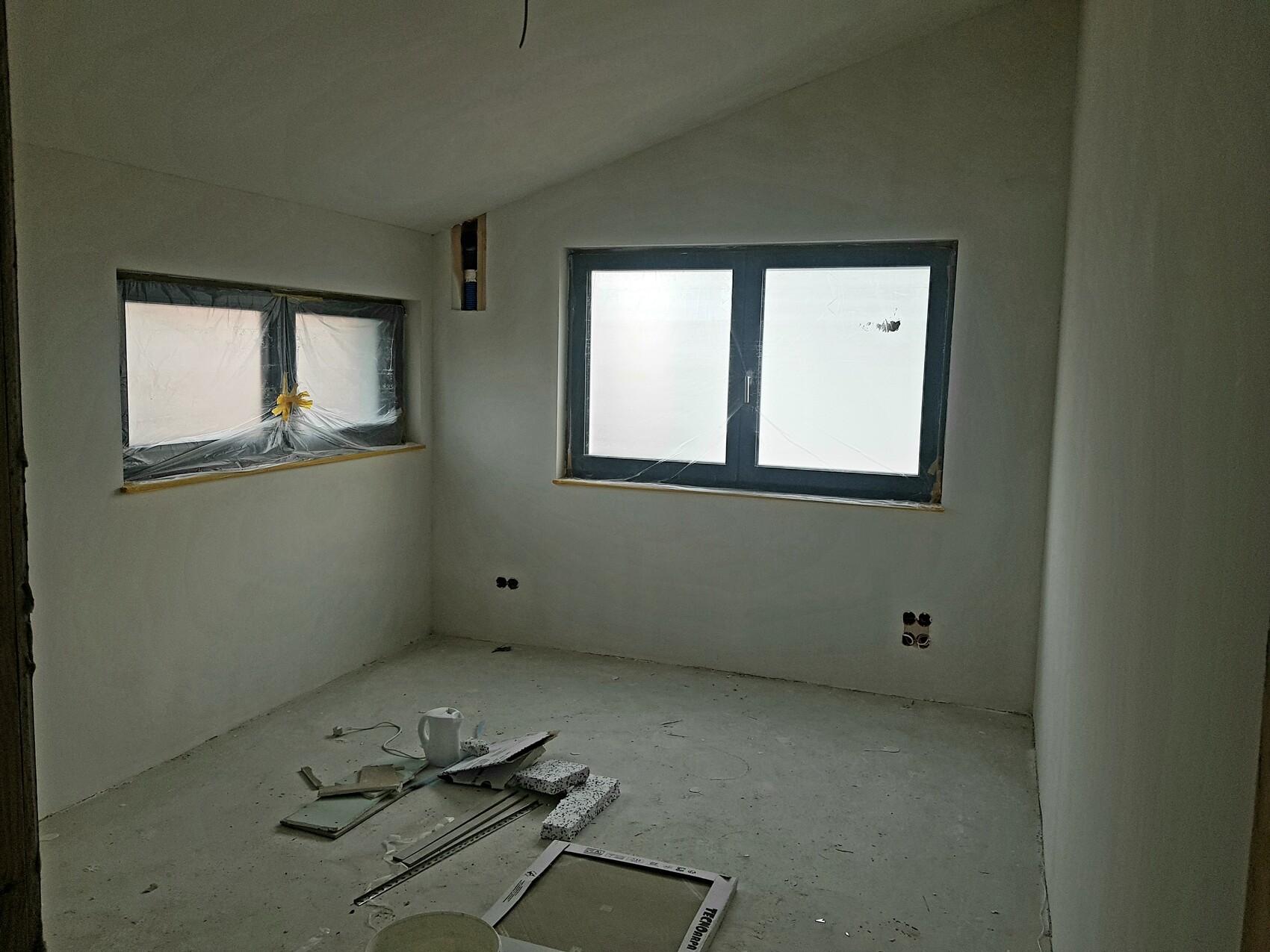 Raum im Bau mit verputzten Wänden, zwei Fenster mit Folie, Werkzeuge und Materialien auf dem Boden.