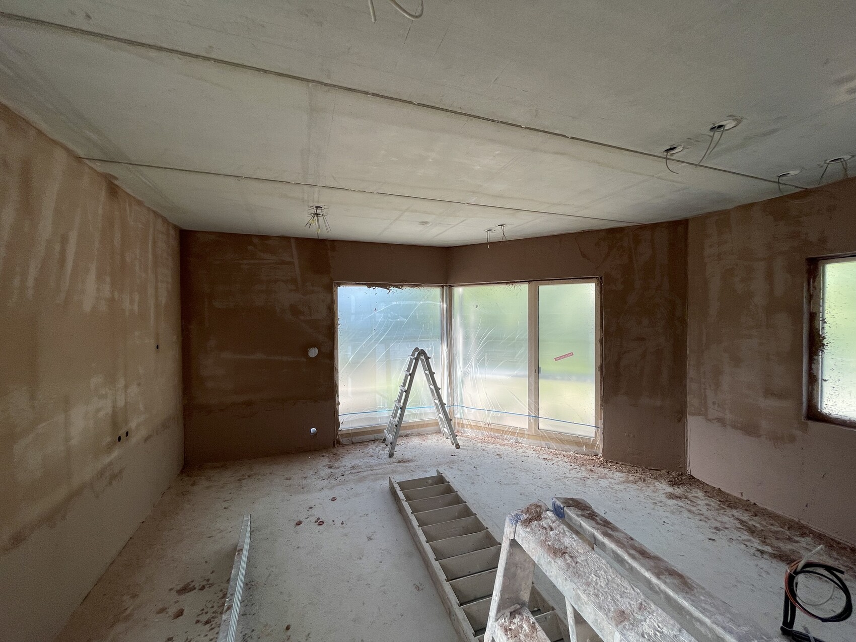 Unfertiges Wohnzimmer-Rohbau: Leiter vor Fenstern, staubige Böden, sichtbare Kabel.