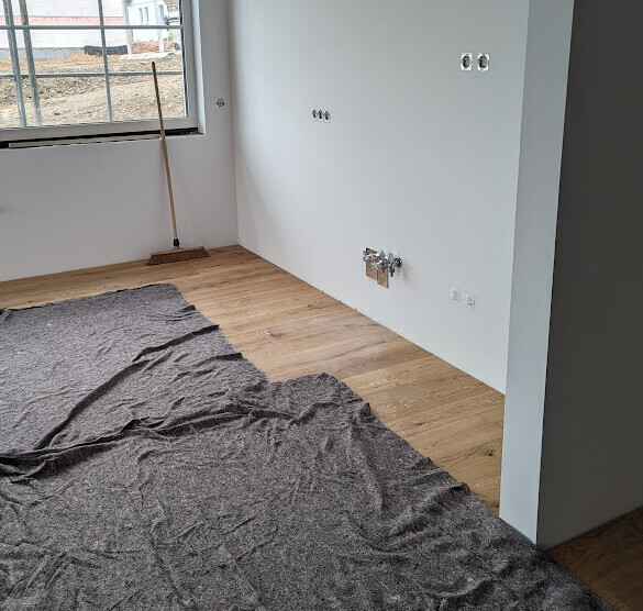 Renovierungsraum: weiße Wände, Holzboden, graue Abdeckung, Besen am Fenster.