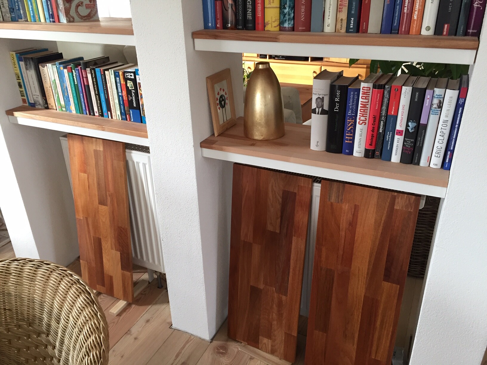 Offenes Bücherregal mit bunten Büchern, goldene Vase, unten Holztüren, Korbstuhl davor.