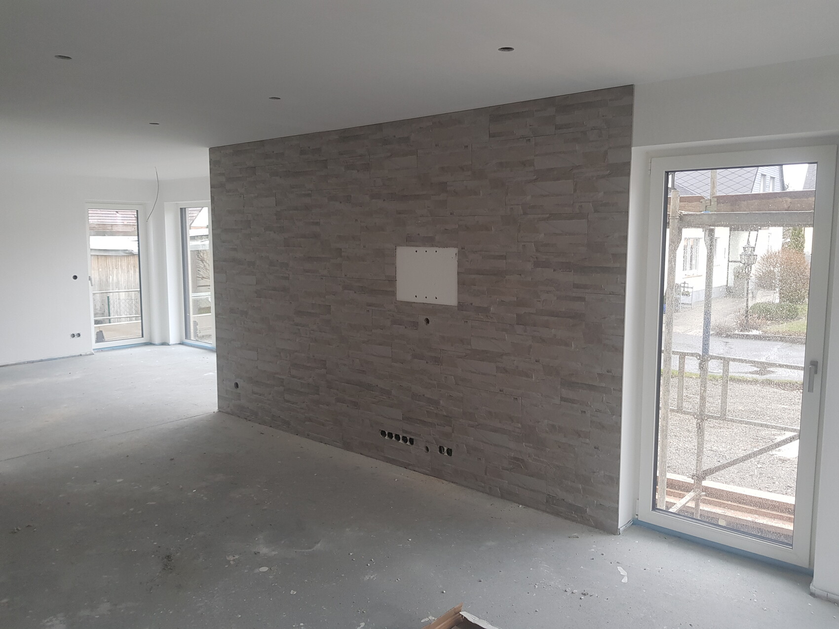 Offenes Wohnzimmer im Rohbau mit grauer Steinwand, großen Glasfenstern und Außentür.